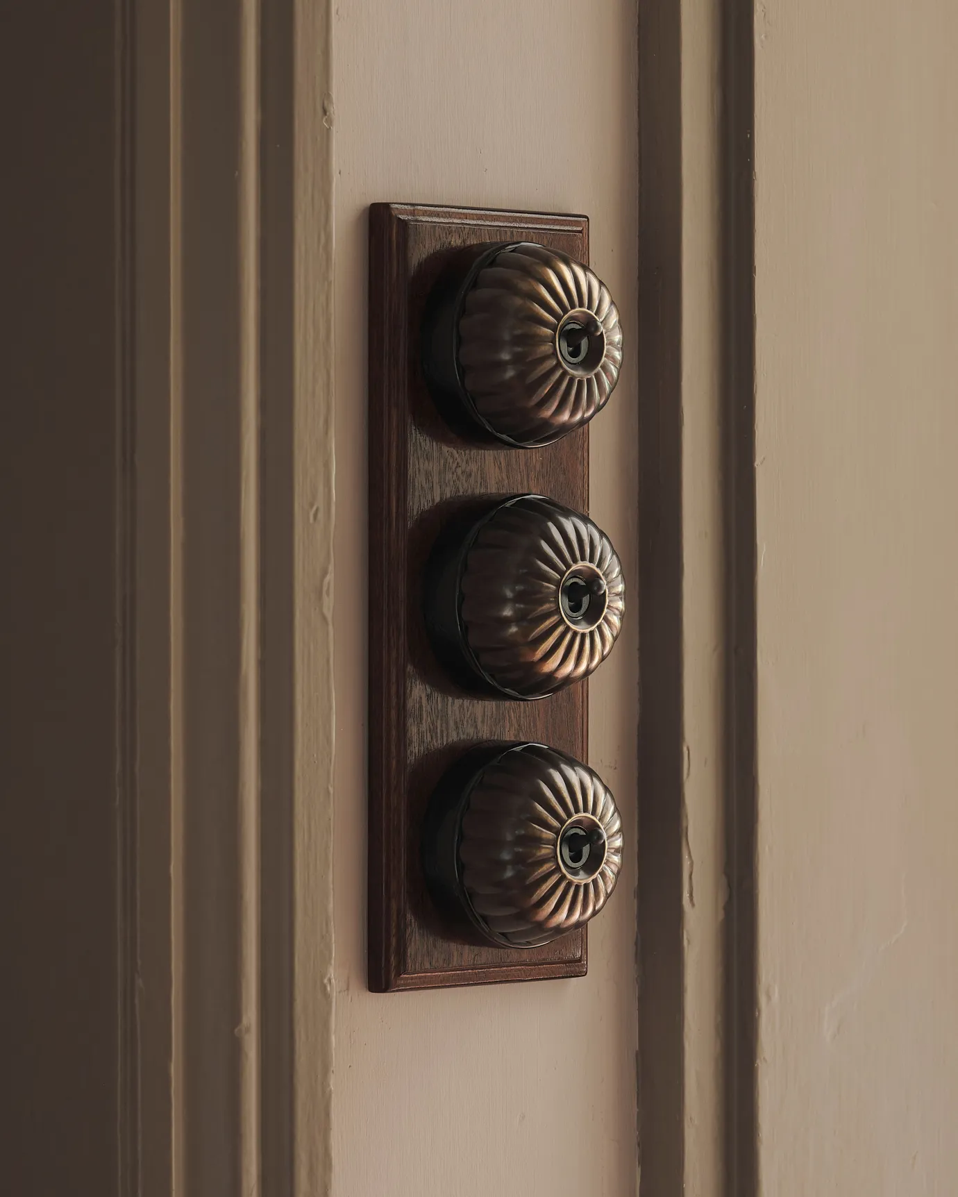 A 3 gang row of Jelly toggle light switches in an Oxidised Brass finish, with black ceramic bases and a Stained Sapele pattress.