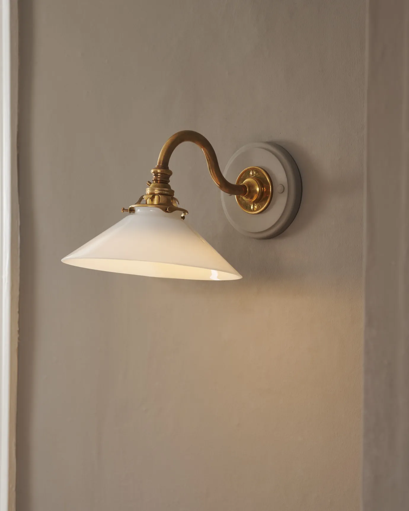 An oblique view of an elegant wall light with a curved brass arm, painted wooden pattress and small milk glass shade. A warm golden light glows through the glass shade.