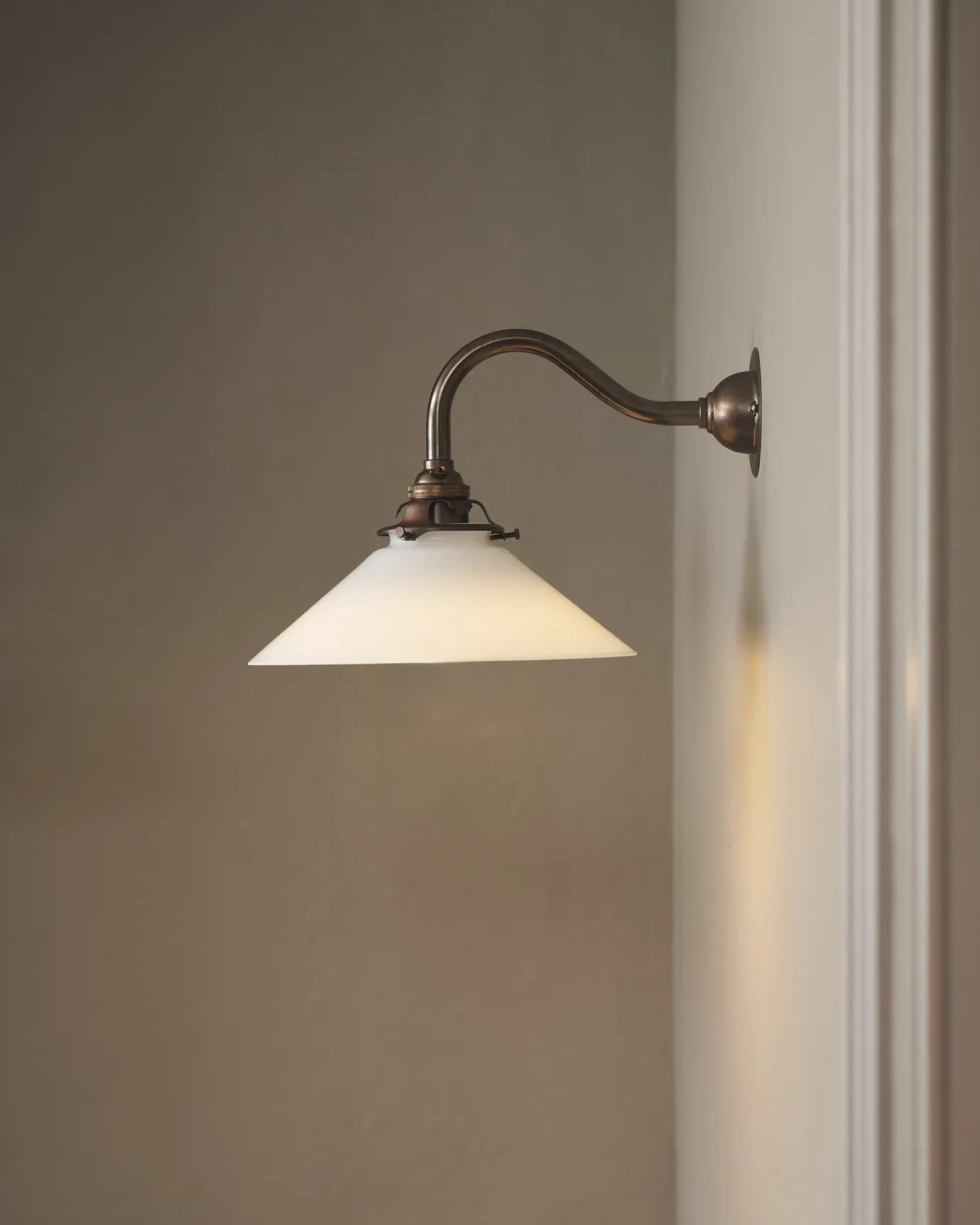 A simple swan neck wall light with an aged brass rose, arm and gallery holding a small illuminated milk glass shade.