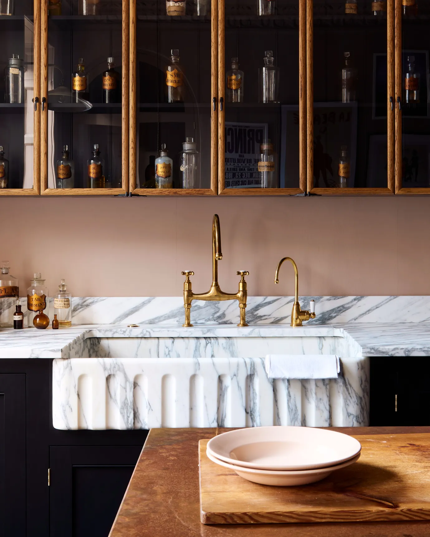 A straight-on view of an extra large single bowl fluted Arabescato marble sink in a black painted Georgian-style kitchen with pink walls and mid-century-style glazed wall cupboards.