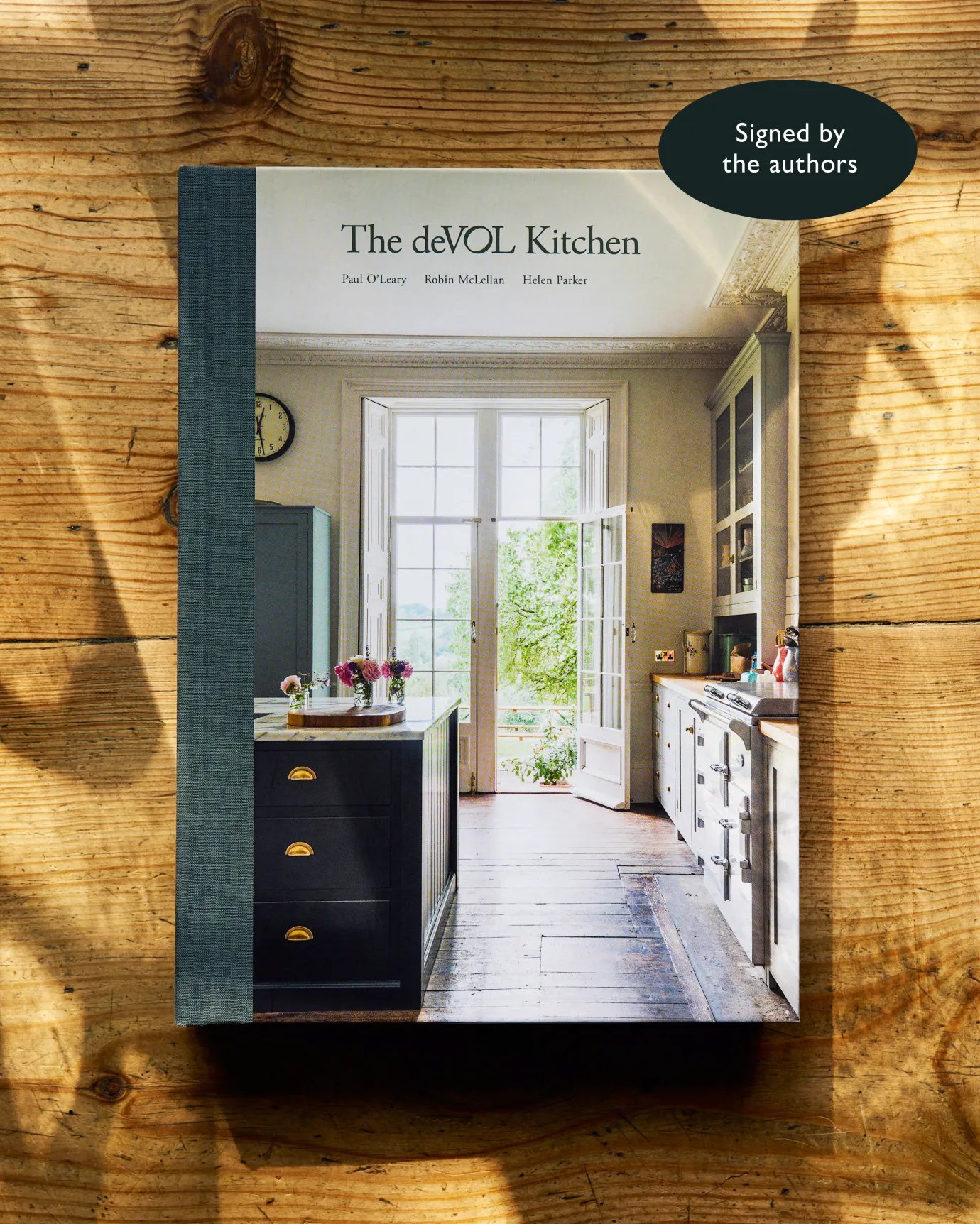 A hardback book on a worn pine table. The title of the book is 'The deVOL Kitchen', it has a blue fabric spine and the cover photograph shows a classic English kitchen with views out over the countryside. An icon indicates that it is a signed copy by the authors.