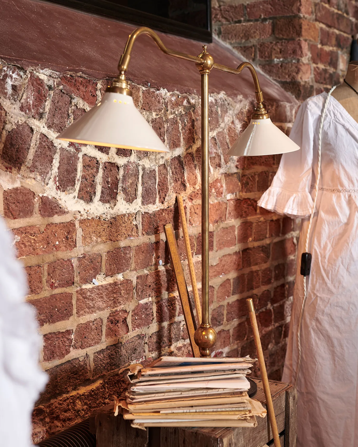 An oblique view of a Victorian-inspired desk lamp with a solid brass foot, stand, two curved arms and two small cream glazed ceramic light shades on a wooden box in front of a red brick wall.