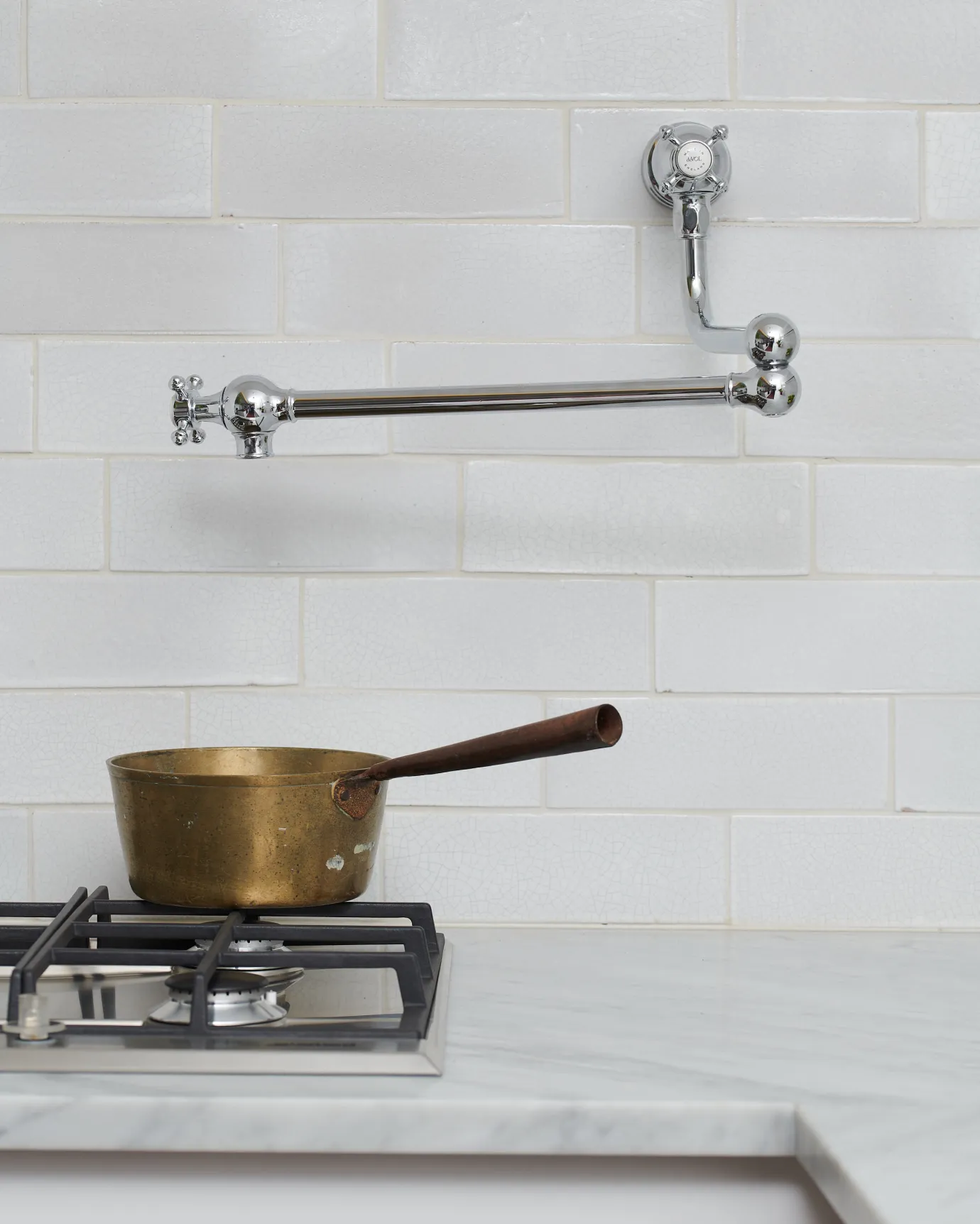 A traditional-style chrome pot filler tap with crosshead handles fitted to a wall of white metro tiles above a gas hob and a vintage copper pan.