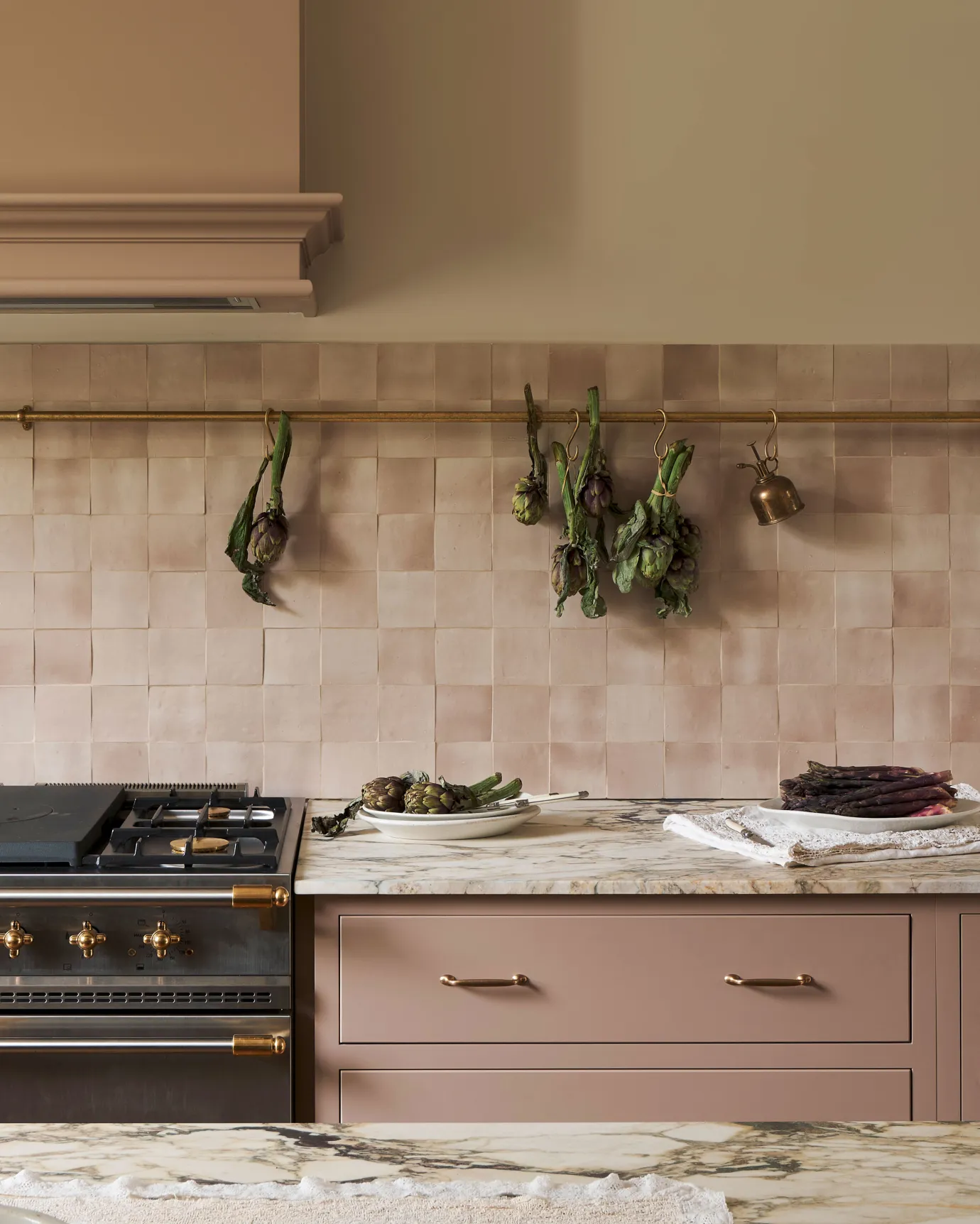 A straight-on view of an aged brass hanging rail fixed to a pink tiled wall above pink painted Shaker-style kitchen cabinets and an Arabescato marble worktop. Several bunches of artichokes hang from brass s hooks along the rail.
