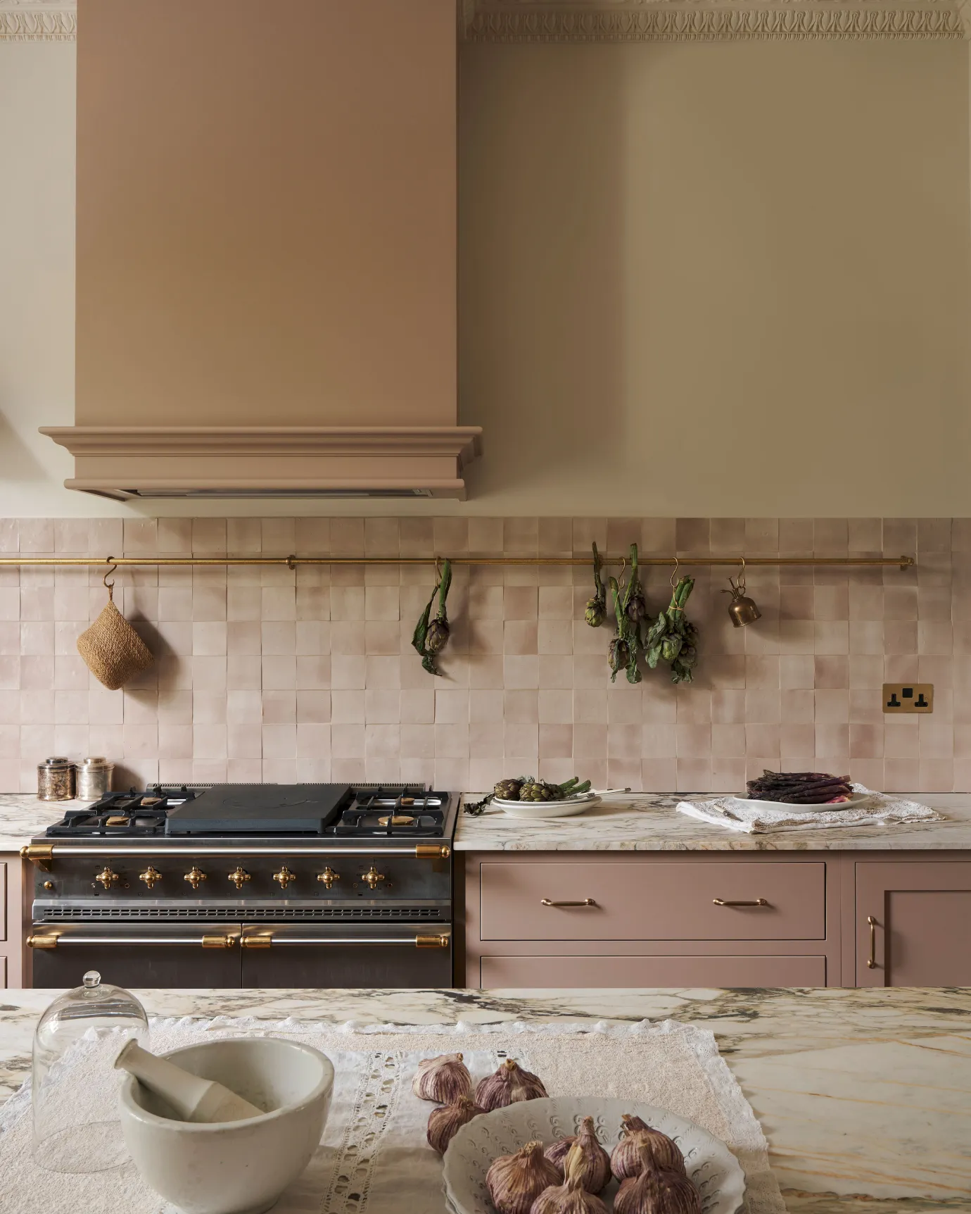 A straight-on view of an aged brass hanging rail fixed to a pink tiled wall above pink painted Shaker-style kitchen cabinets and an Arabescato marble worktop. Several bunches of artichokes hang from brass s hooks along the rail.