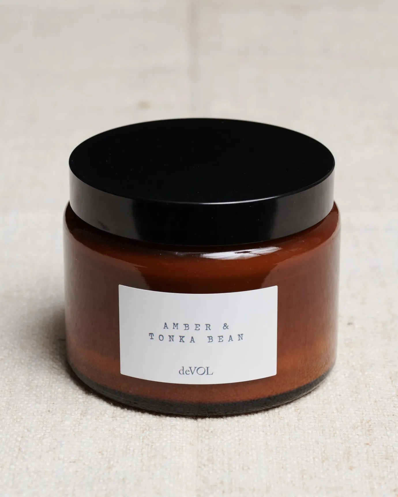 A large scented candle in a brown glass jar with a black lid, the label reads 'Amber & Tonka Bean, deVOL'.