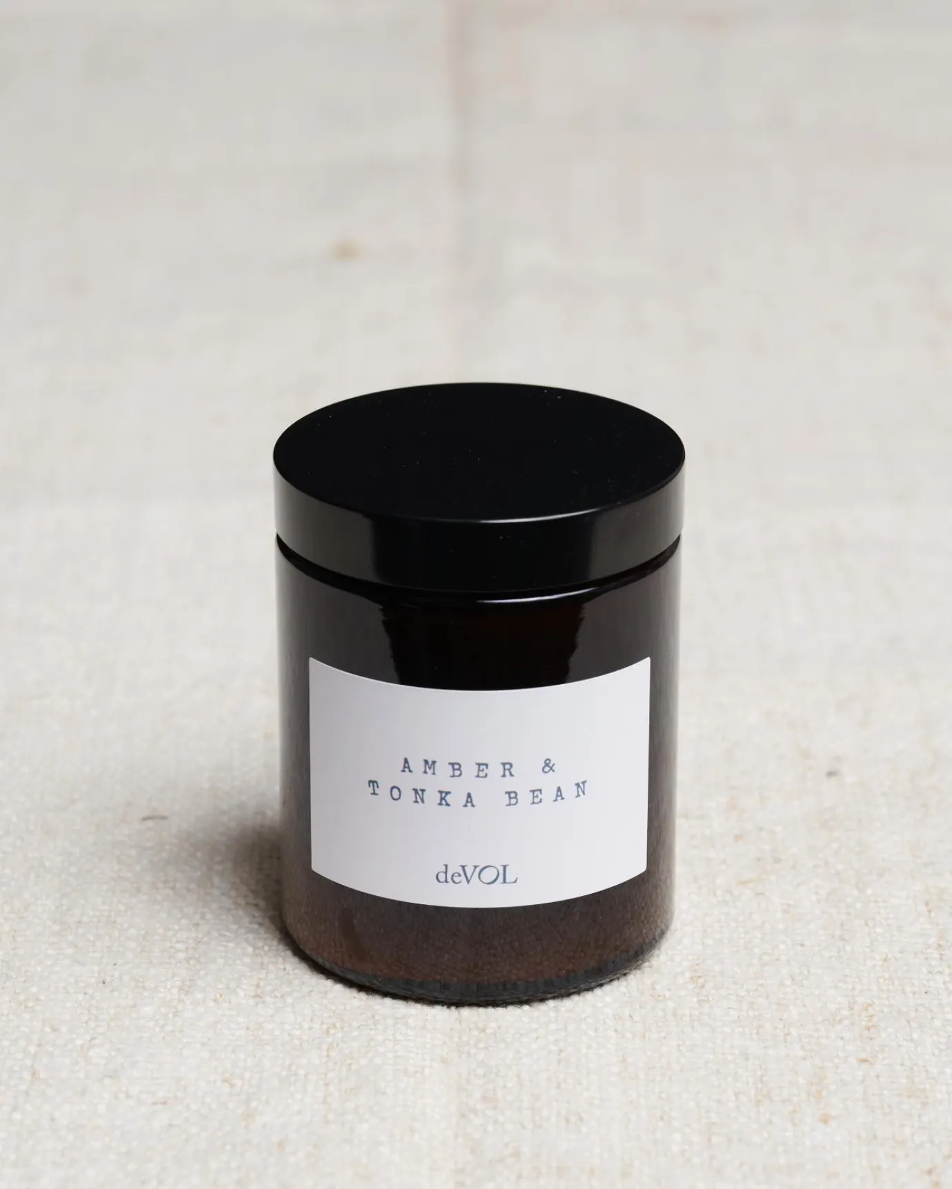 A medium scented candle in a brown glass jar with a black lid, the label reads 'Amber & Tonka Bean, deVOL'.