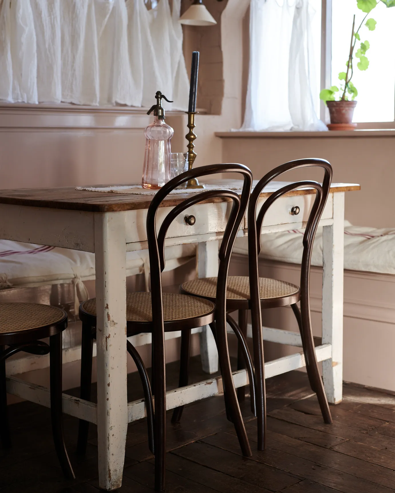 An oblique view of two café-style bentwood chairs with dark stained frames and light woven rattan seat mesh tucked into a vintage pine table.