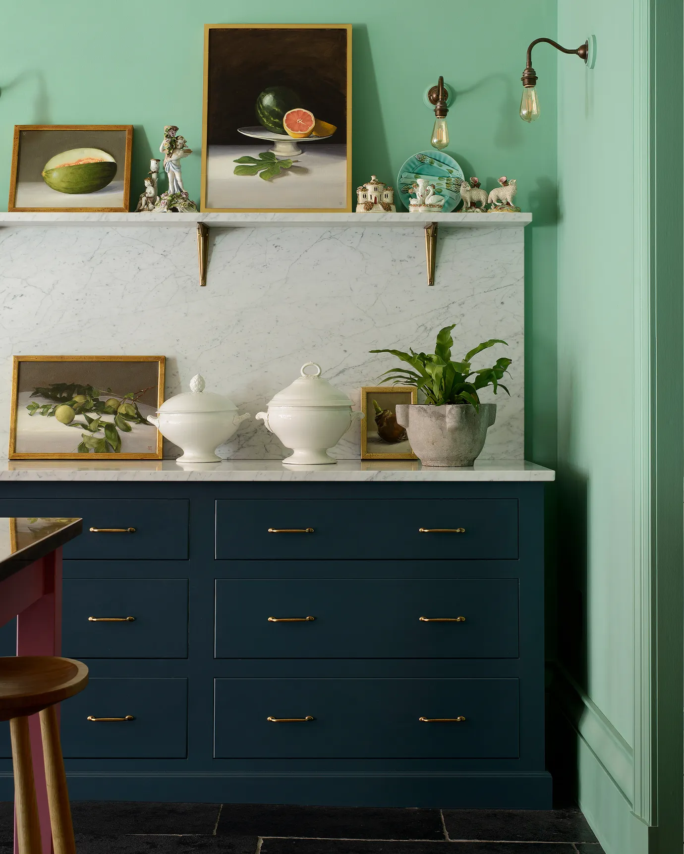A straight-on view of Georgian-style kitchen drawers painted in a royal blue, with brass hardware, a Carrara marble worktop and splashback, vintage bric-a-brac and light arsenic green walls.