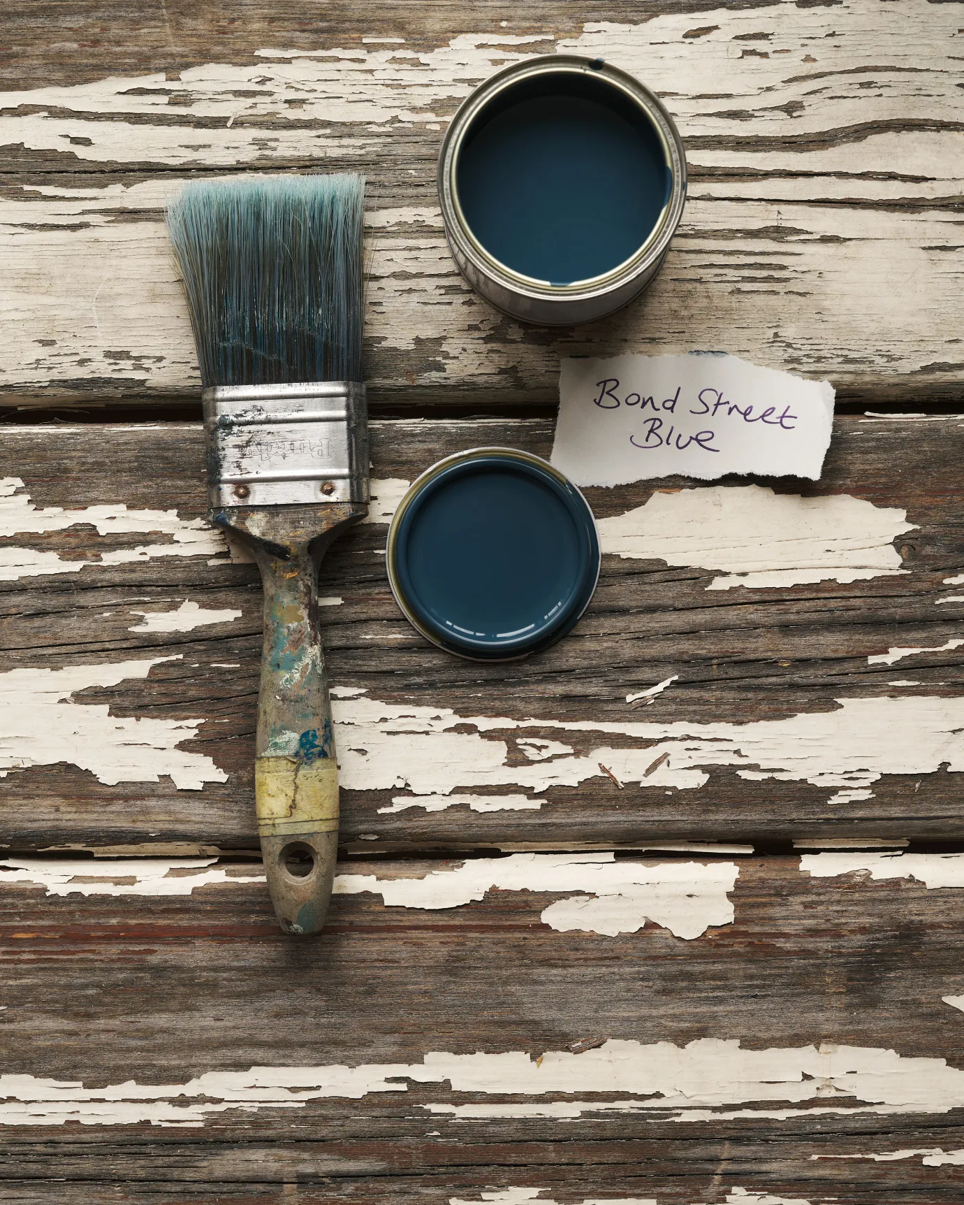 An overhead view of an open paint pot and a worn brush on a rustic tabletop. The royal blue colour inside is labelled 'Bond Street Blue'.