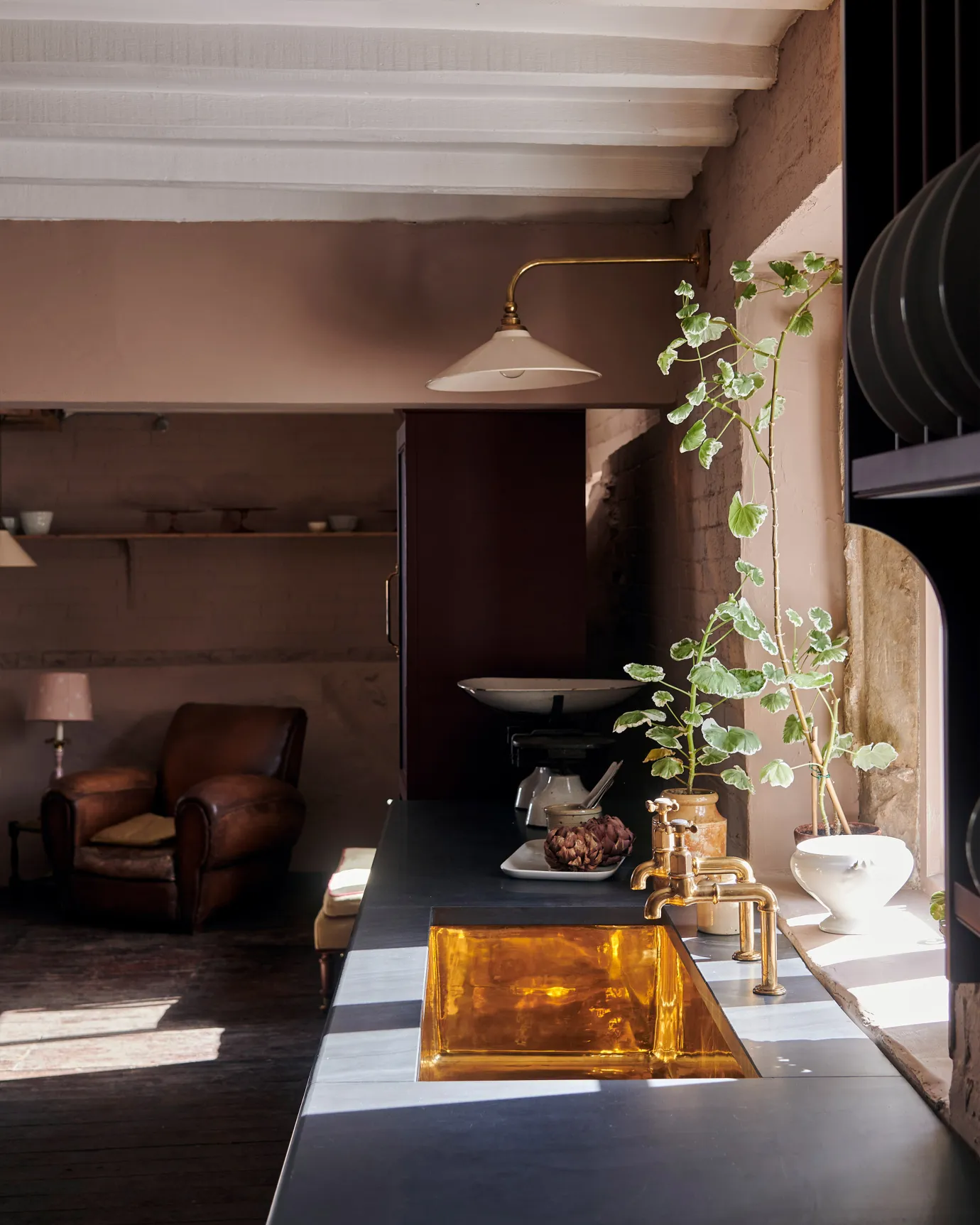 Looking across a slate kitchen worktop with a large undermounted polished brass sink with aged brass bibcock taps and a tall geranium plant.