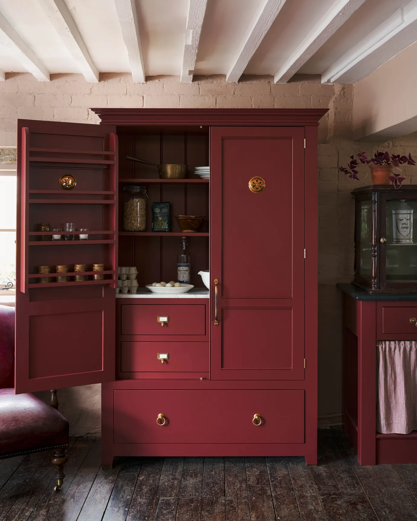 A red painted Georgian-style pantry cupboard with brass hardware and Victorian-inspired circular brass ventilation grilles fitted to each door. One door is open to reveal the decorative brass backplate of the ventilation grille.