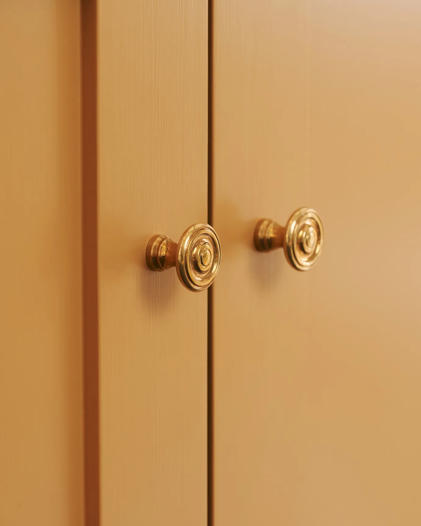 A close-up oblique view of a pair of small Victorian-inspired doorknobs in a warm burnished brass finish, fitted to a yellow painted cupboard doors.