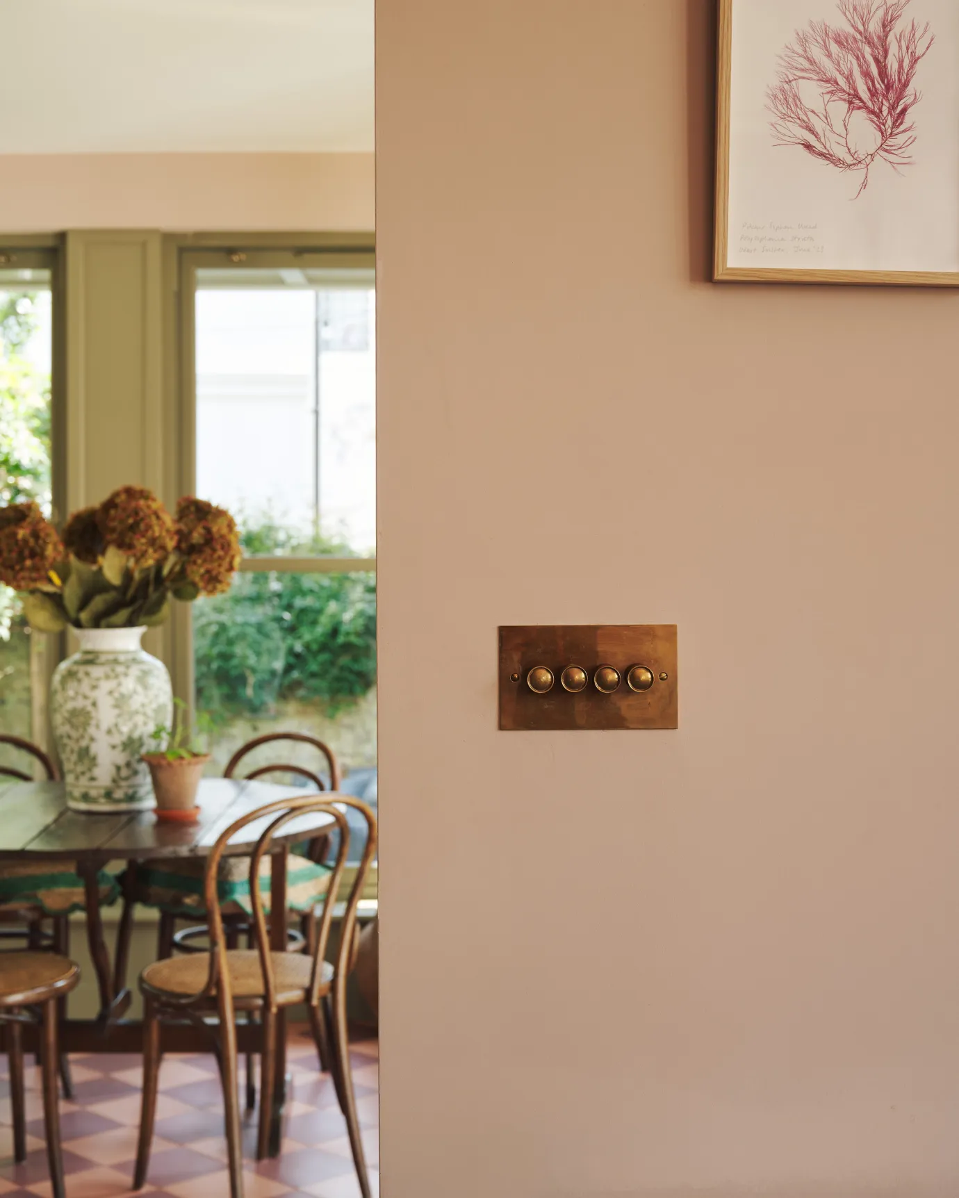 A straight-on view of a 4 gang classic dimmer switch in a warm burnished brass finish on soft pink walls, a café-style table and chairs with a vase full of pink hydrangeas can be seen in the background.