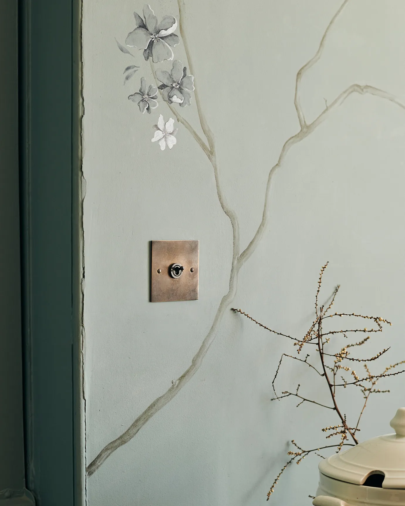 An oblique view of a 1 gang classic toggle switch in a dark oxidised brass finish on a duck-egg blue wall with a delicate handpainted mural of winding branches.