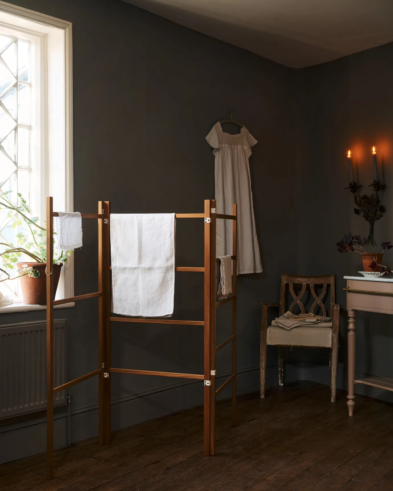 The corner of a dark room with a leaded Victorian window and wooden floorboards, a linen dress hanging from a rail behind a wooden clothes horse draped in vintage linens.