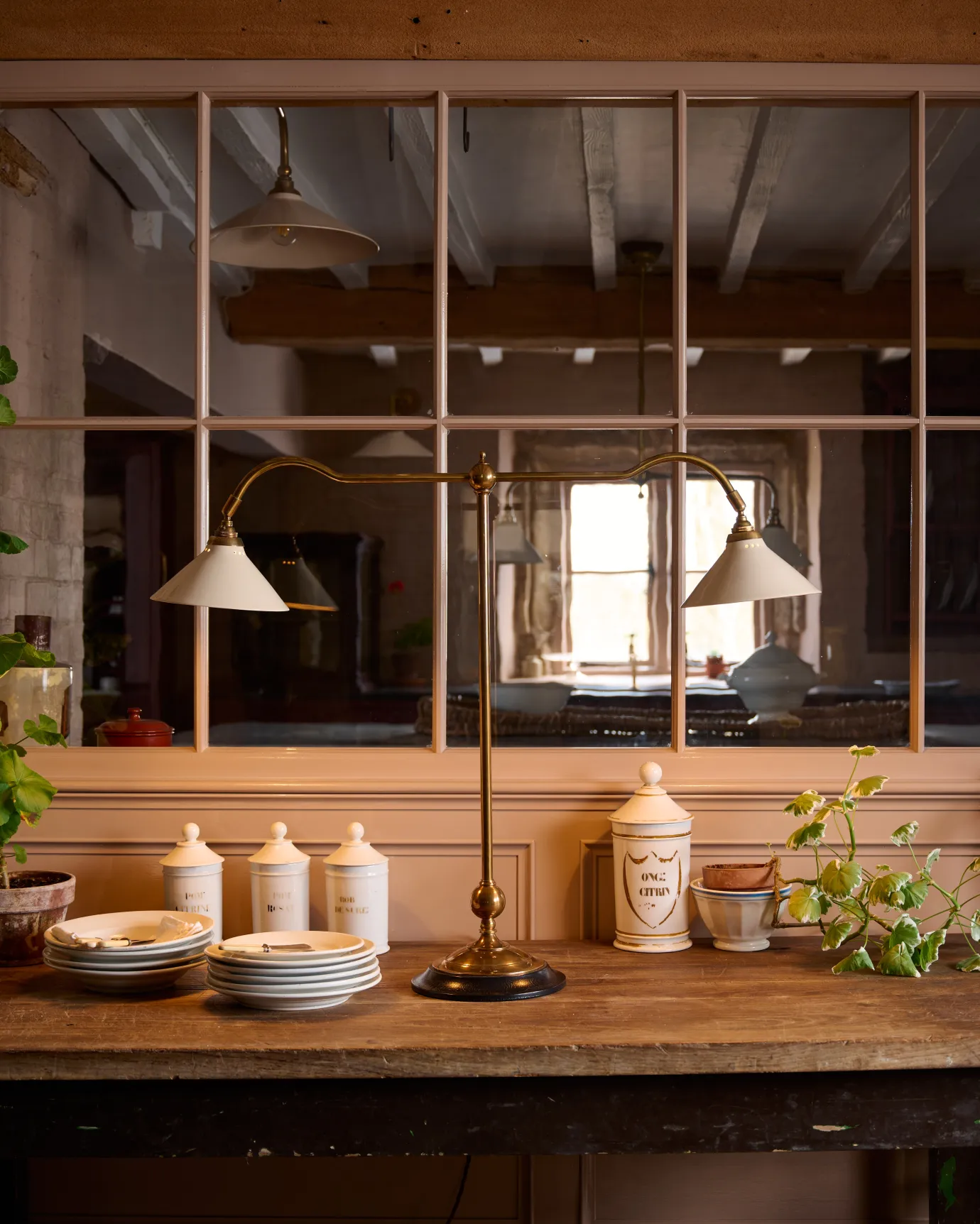 A straight-on view of a Victorian-inspired desk lamp with a solid brass foot, stand, two curved arms and two small cream glazed ceramic light shades on a table in front of a glazed partition.