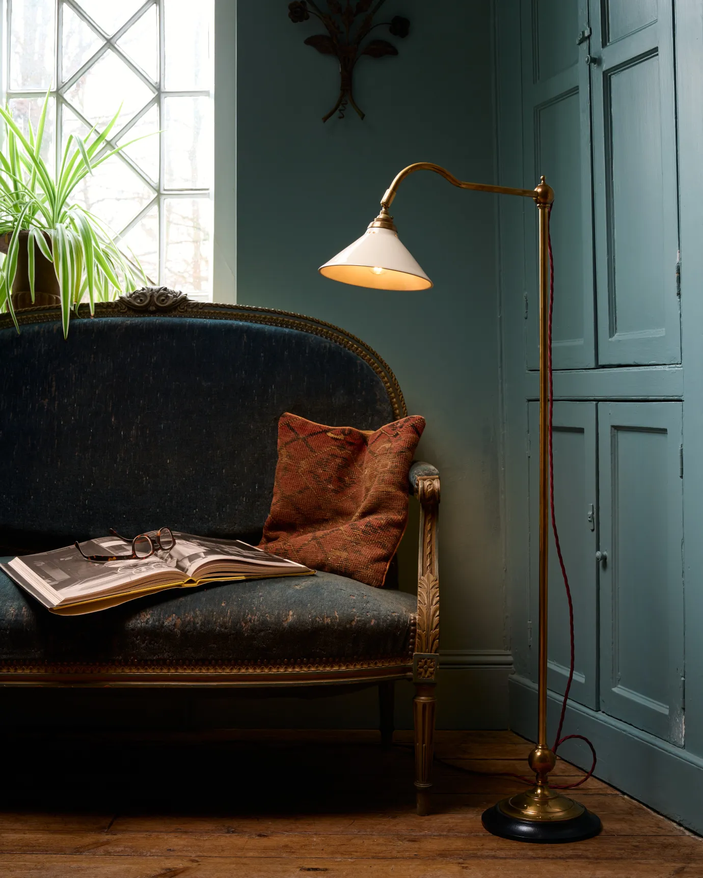 A straight-on view of a Victorian-inspired floor lamp with a solid brass foot, stand, curved arm, dark hardwood base and a small cream glazed ceramic light shade in a living room with blue walls, dark sofa and leaded windows.