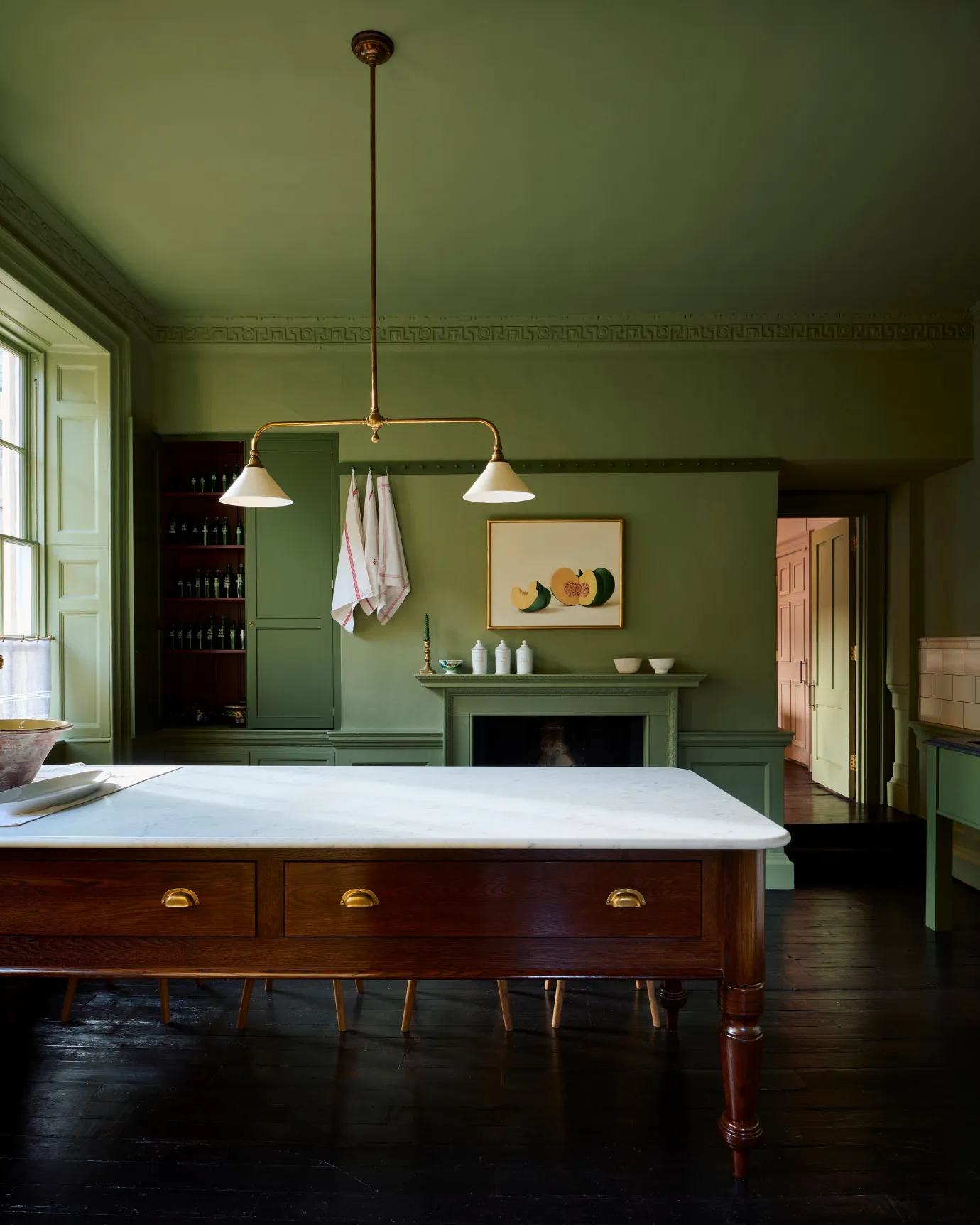 An atmospheric room with dark green walls and ceiling, black floorboards and a Victorian-inspired chandelier light with brass arms and small unglazed porcelain shades over a large oak prep table with a Carrara marble worktop.
