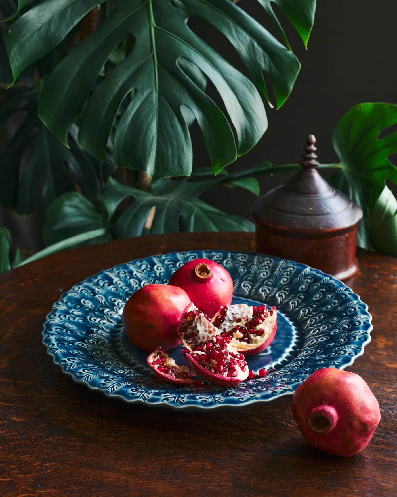 Bright ruby pomegranates on a large handmade bohemian-style blue ceramic platter with a scalloped rim and delicate impressions and surface patterns. The colours are complimented by a dark wood round table and a large green monstera plant in the background.