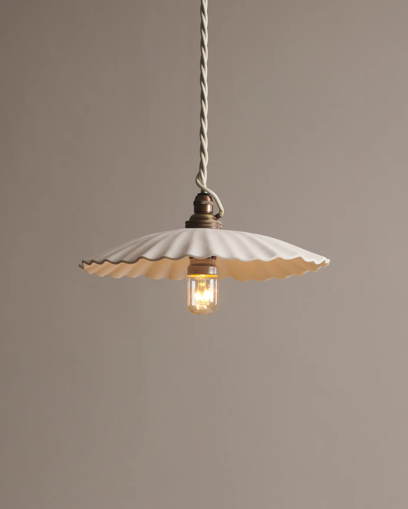 A large narrow pleat porcelain pendant light with an unglazed finish, an aged brass B22 lampholder and a white twisted fabric flex cord.