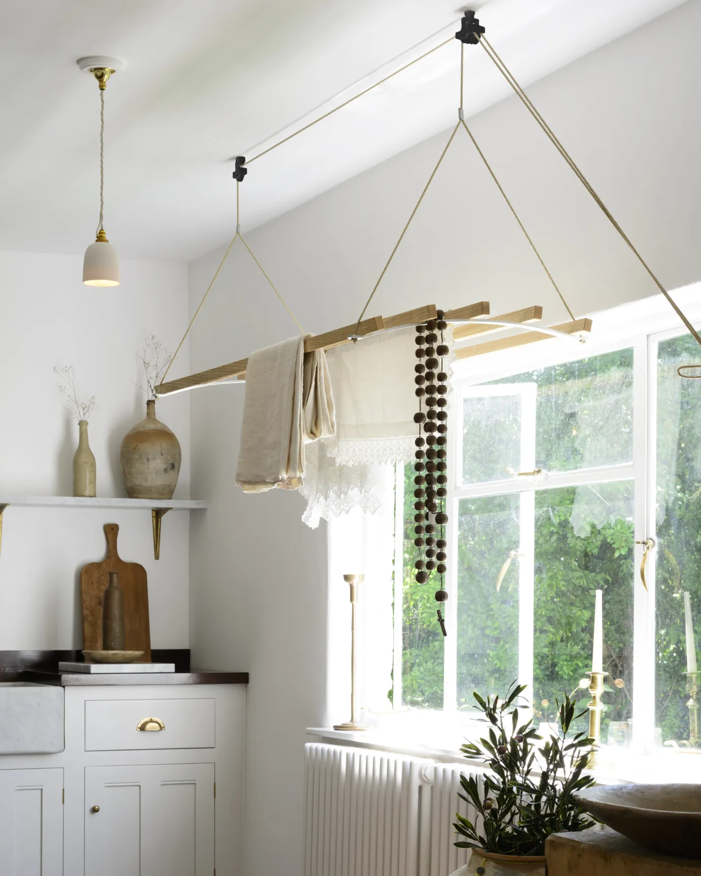 An oblique view of a laundry maid with wooden slats and aluminium brackets, hanging from the ceiling in a Mediterranean-inspired room with white walls, terracotta flooring and rustic styling.