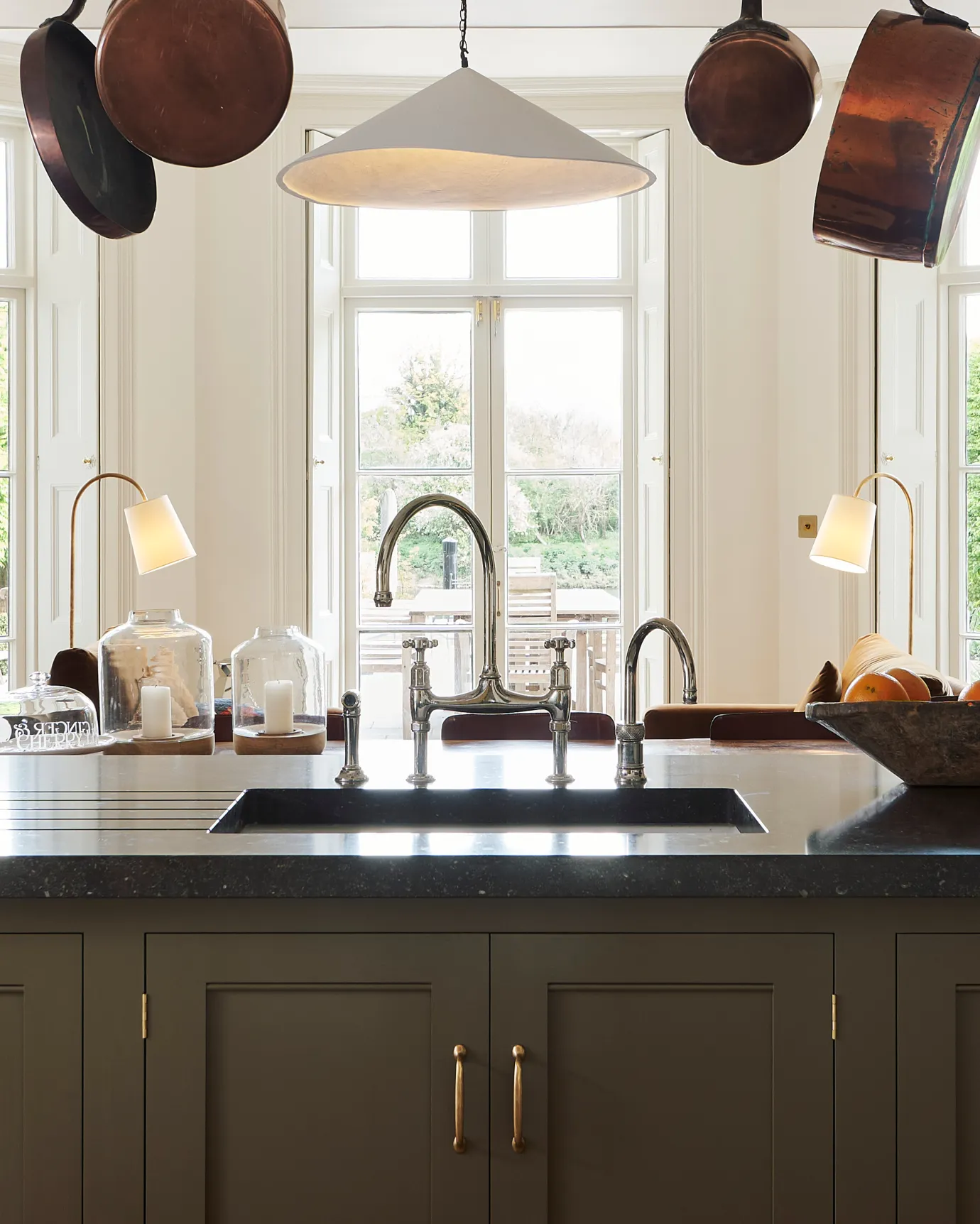 An oblique view of a traditional-style nickel mixer tap with crosshead handles in a simple cream-coloured Shaker kitchen with louvred shutters behind.