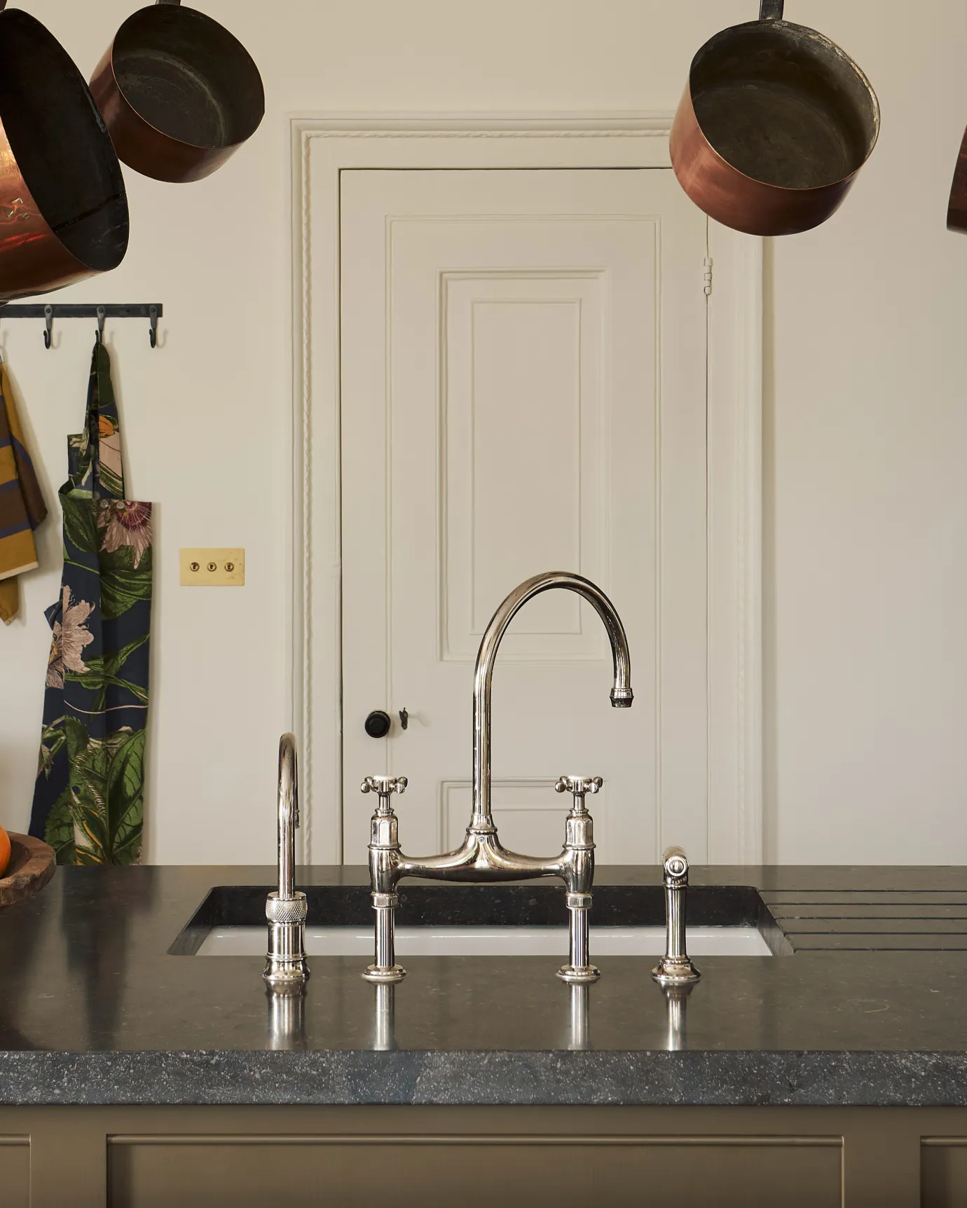 A straight-on view of a traditional-style nickel mixer tap with crosshead handles in a simple cream-coloured Shaker kitchen with louvred shutters behind.