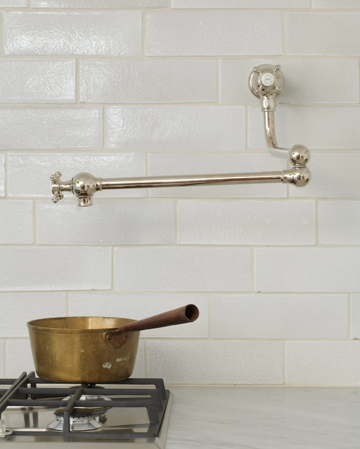 A traditional-style nickel pot filler tap with crosshead handles fitted to a wall of white metro tiles above a gas hob and a vintage copper pan.