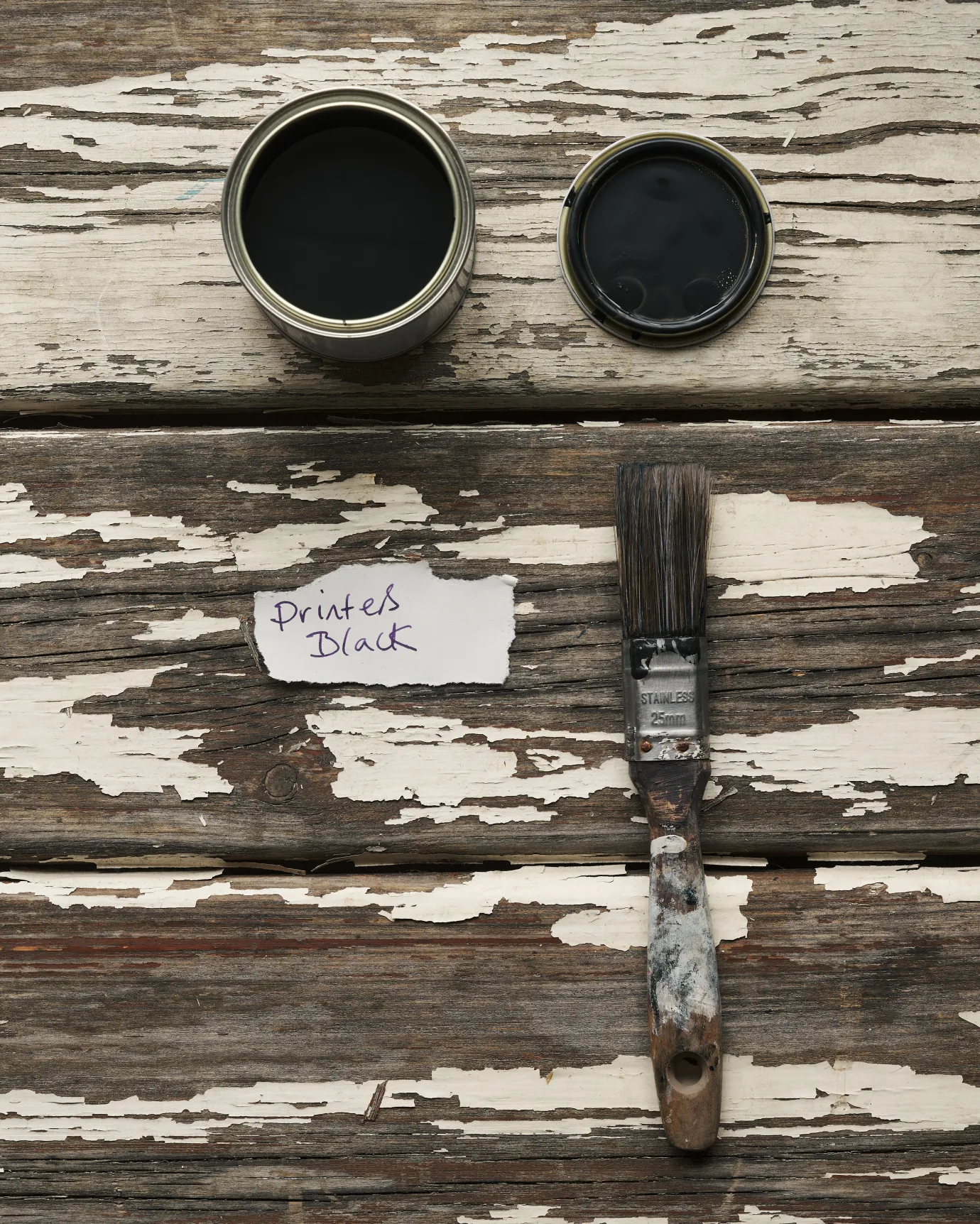 An overhead view of an open paint pot and a worn brush on a rustic tabletop. The black colour inside is labelled 'Printer's Black'.