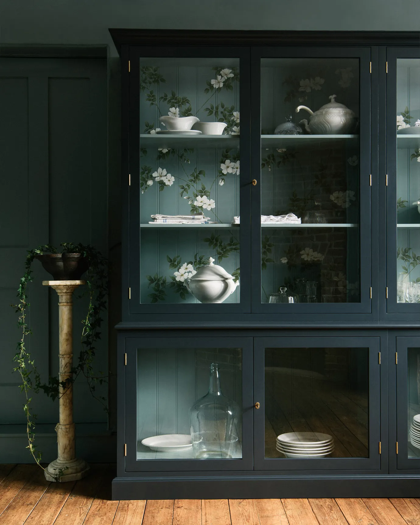 A tall museum-style display cabinet with glazed doors, painted a dark blue outside with a duck-egg blue interior and painted flowing white dog-roses on the tongue and groove back panel.