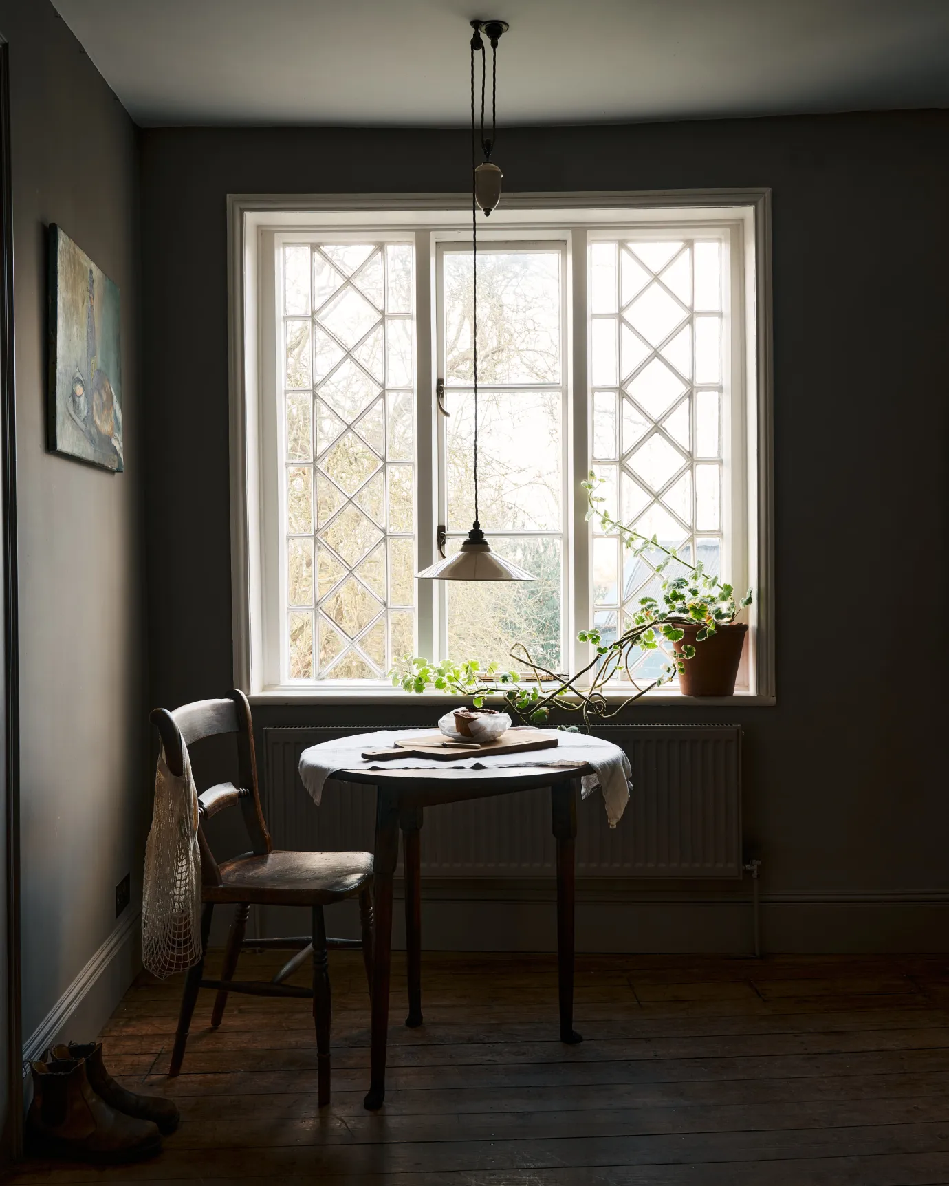 An oblique view of a rise and fall light with a small cream glazed ceramic shade, aged brass fittings and brown twisted fabric flex cord hanging in a dark room by a large Victorian leaded window.