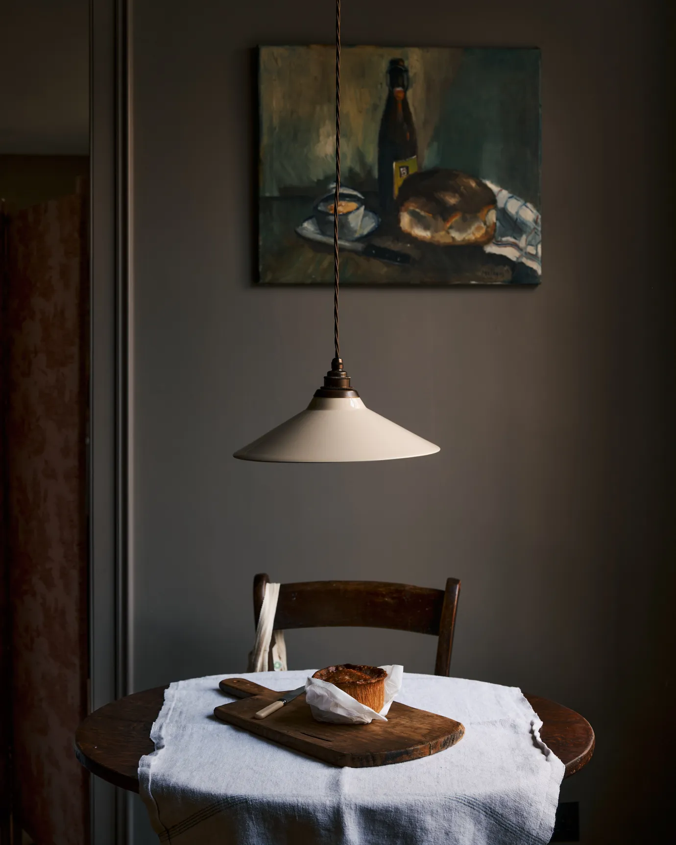 A straight-on view of a rise and fall light with a small cream glazed ceramic shade, aged brass fittings and brown twisted fabric flex cord hanging in a dark room over a small round wooden table with a linen tablecloth and fresh bread.