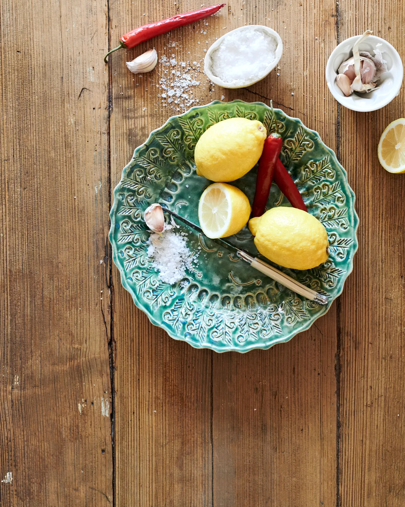 An overhead view of a round handmade bohemian-style green ceramic platter with delicate impressions and surface patterns on a rustic pine tabletop with vintage silverware, lemons, salt, chillis and garlic.