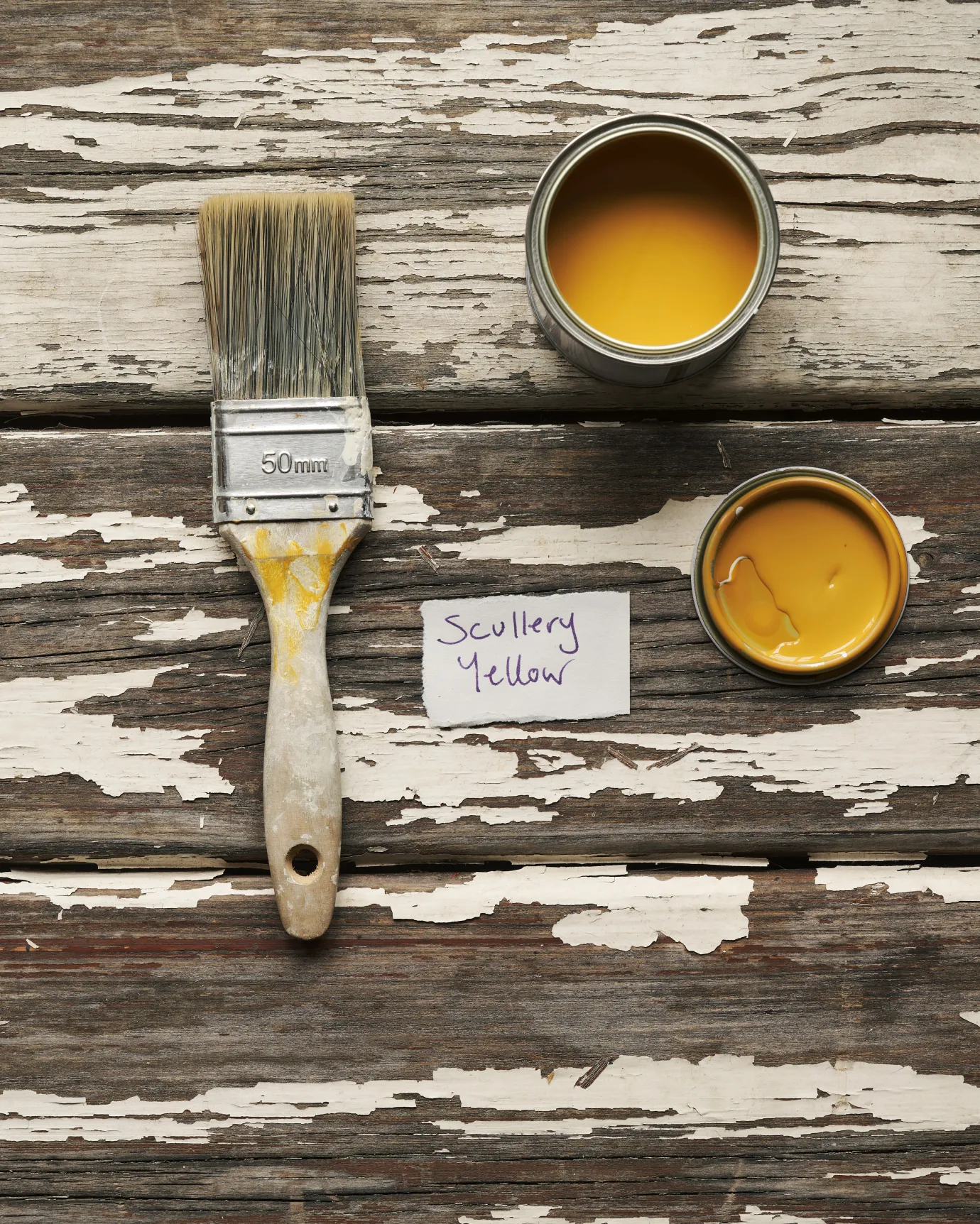 An overhead view of an open paint pot and a worn brush on a rustic tabletop. The warm yellow colour inside is labelled 'Scullery Yellow'.