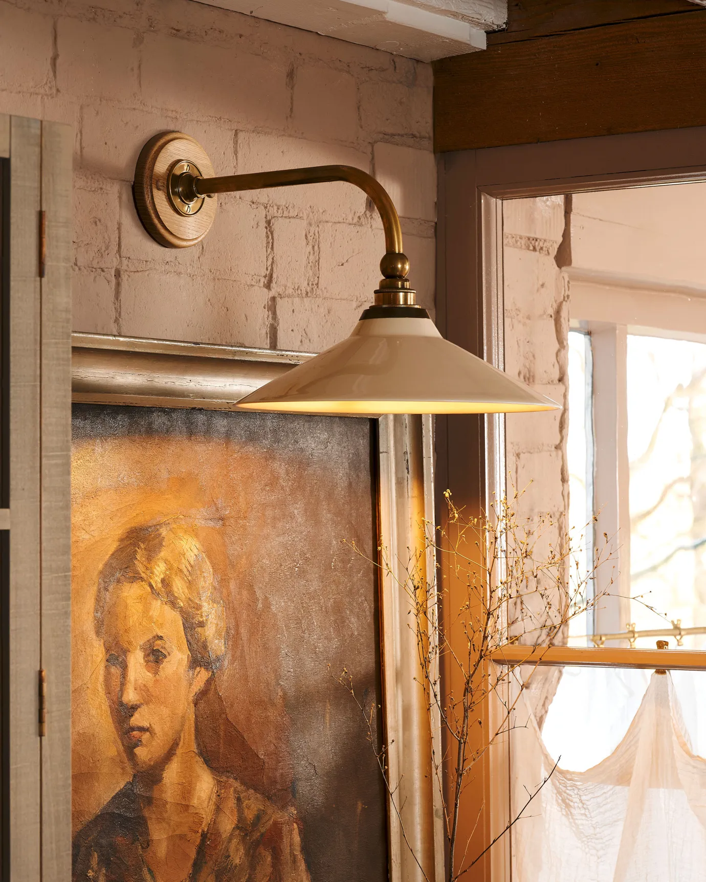 A close-up oblique view of a small Victorian-inspired wall light with a brass arm and a cream glazed ceramic shade fixed to a pink brick wall above an antique portrait painting.