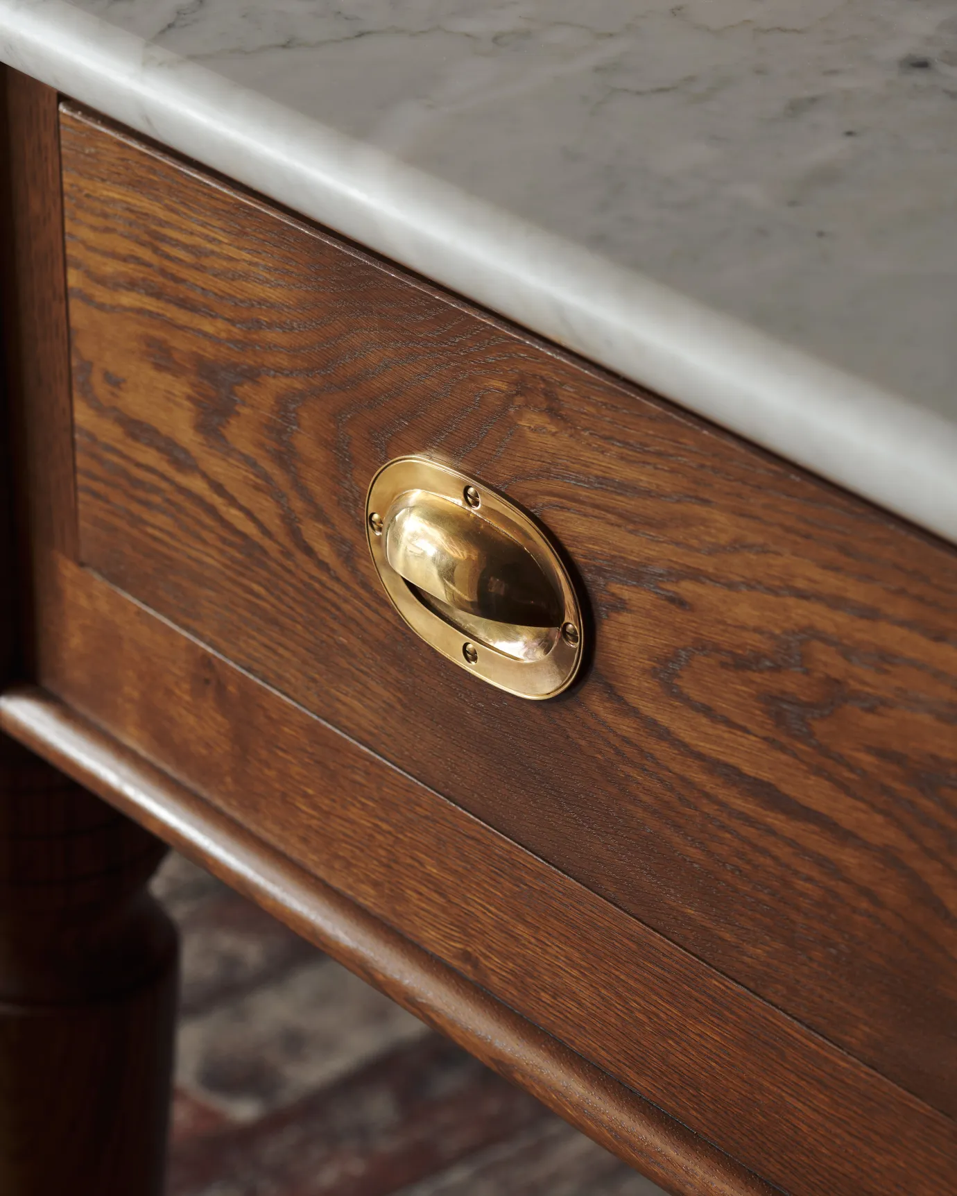 An oblique view of a Victorian-inspired oval-shaped cup handle with an authentic and warm burnished brass finish and a recessed backplate, fitted to a dark stained oak drawer front.