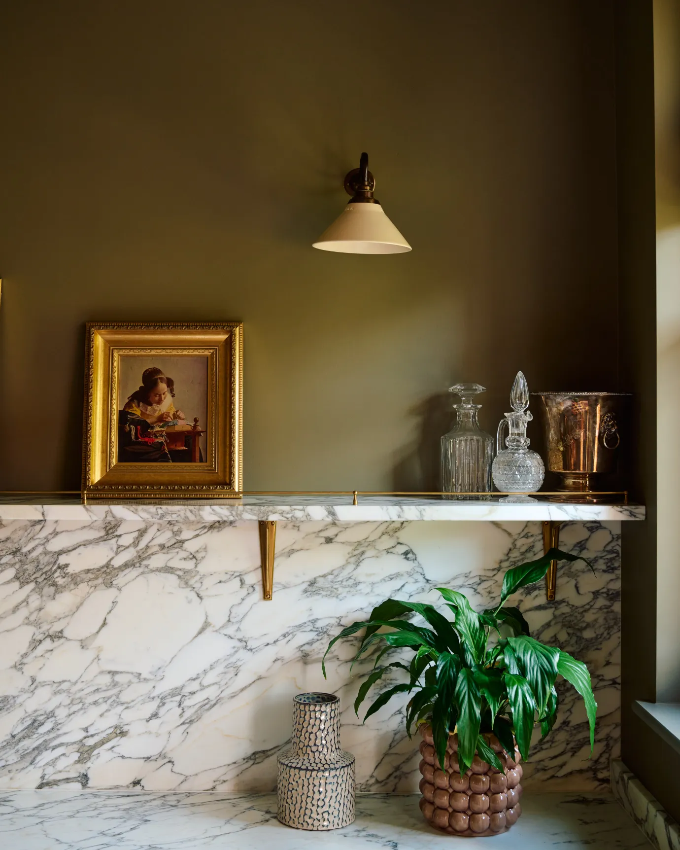A straight-on view of a small wall light with an aged brass swan neck arm and a small cream glazed ceramic shade, fixed to a dark green wall above an Arabescato marble splashback and shelf, decorated with antique oil paintings in gilded frames.