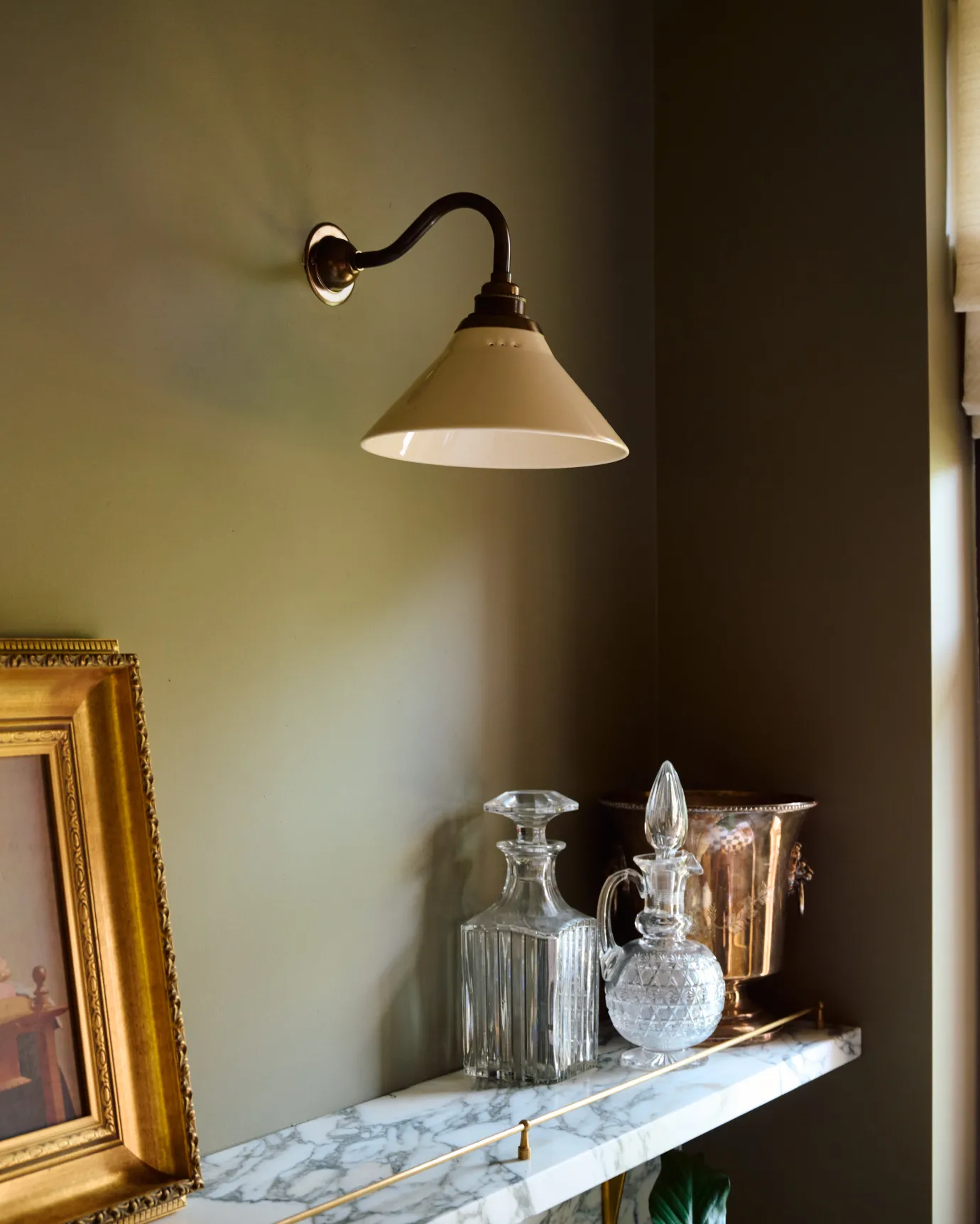 An oblique view of a small wall light with an aged brass swan neck arm and a small cream glazed ceramic shade, fixed to a dark green wall above an Arabescato marble shelf with glass bottles.