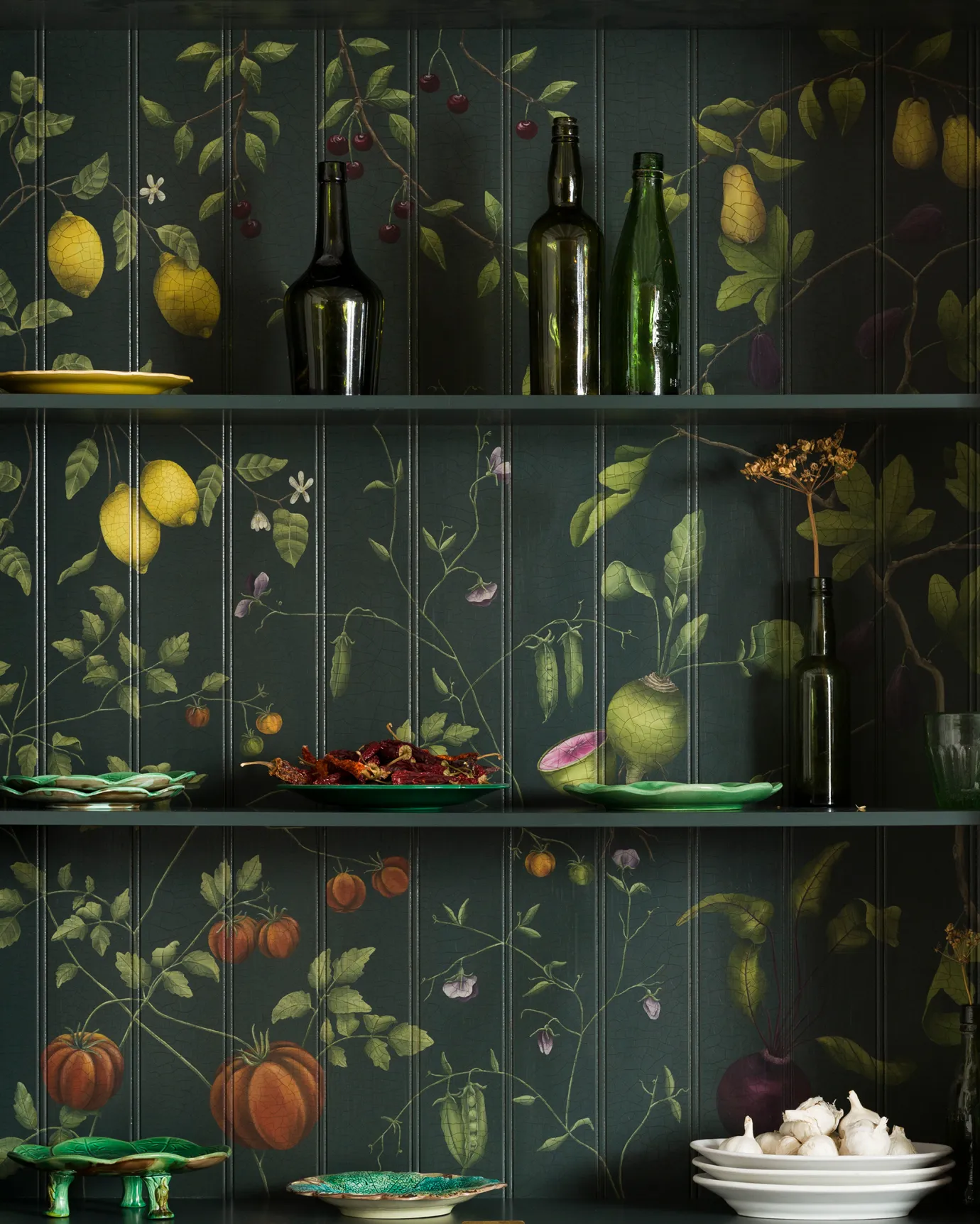 The interior of a display cabinet with a dark green interior and a traditional painted botanical design on the tongue and groove back panel. The shelves are dotted with vintage crockery and green glass bottles.