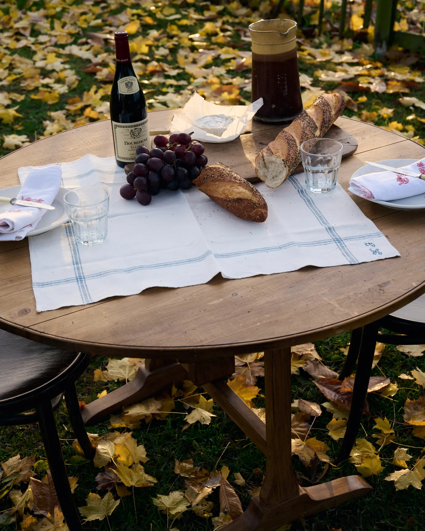A close-up view of a round pine dining table outdoors with a small vintage linen tablecloth and a spread of red wine, cheese, bread and grapes.