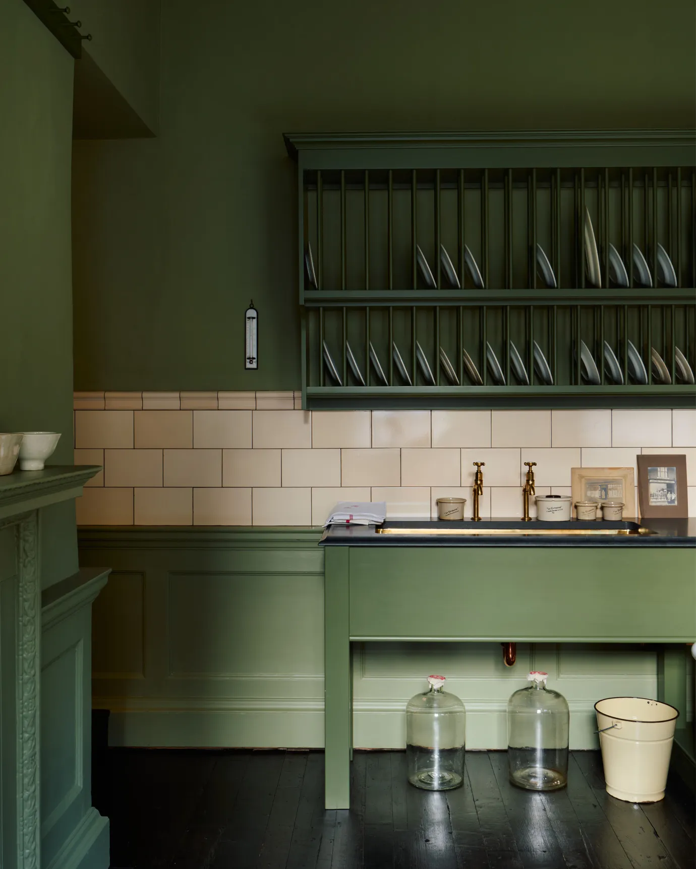 A straight-on view of a mossy green painted Victorian-style sink cupboard with a polished brass sink, aged brass bibcock taps and cream subway wall tiles below a traditional wall-mounted plate rack.